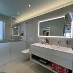 Modernes Badezimmer mit Glaswand, beleuchtetem Spiegel und eleganter Waschbeckenanordnung. Minimalistisches Design mit neutralen Farben.