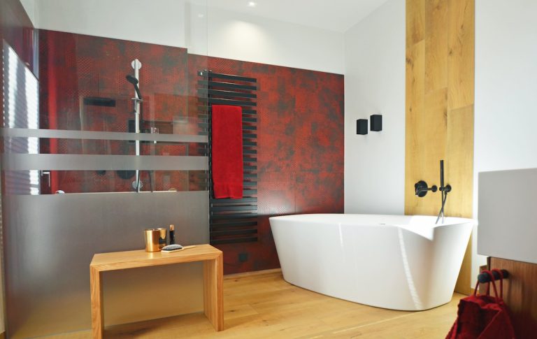 Modernes Badezimmer mit freistehender weißer Badewanne, roter Akzentwand mit dunklem Muster und begehbarer Dusche mit Milchglaswand. Schwarze Armaturen und ein hölzerner Hocker ergänzen das elegante Design. Das Badezimmer wurde vom Kölner Unternehmen Holthausen Exklusiv modernisiert.