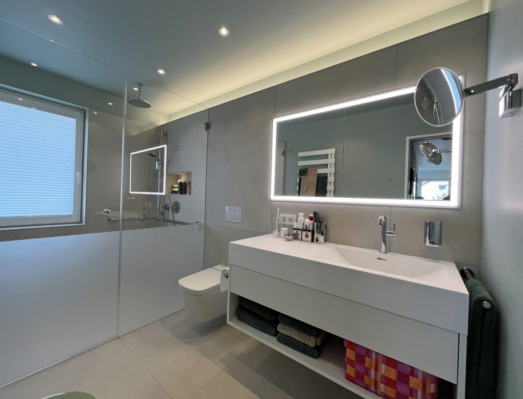 Modernes Badezimmer mit Glaswand, beleuchtetem Spiegel und eleganter Waschbeckenanordnung. Minimalistisches Design mit neutralen Farben.