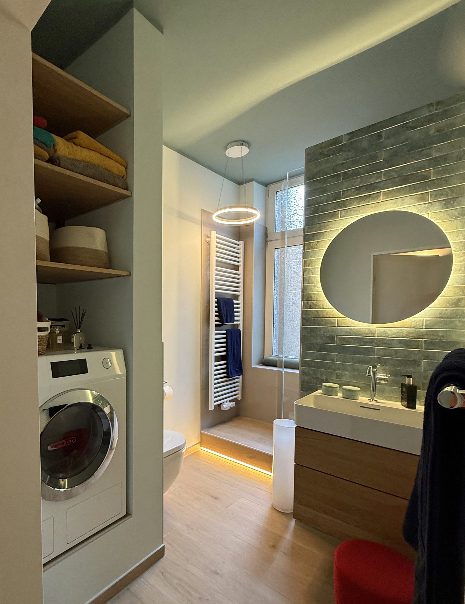 Modernes Badezimmer mit Waschmaschine, runder Spiegelleuchte und Holzwaschtisch. Wand mit grünen Fliesen und Handtuchhalter.